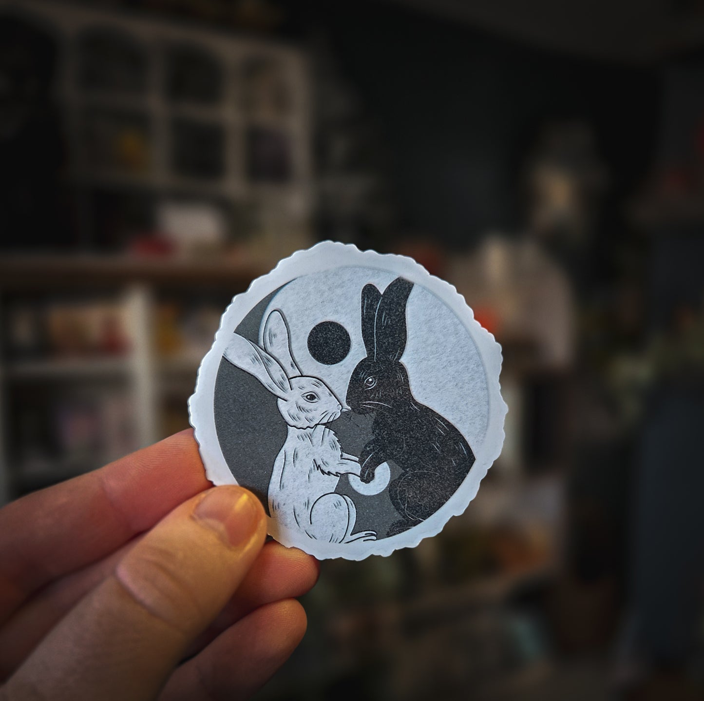 Hand holding a round sticker with black and white illustrations of two rabbits against a dark background.